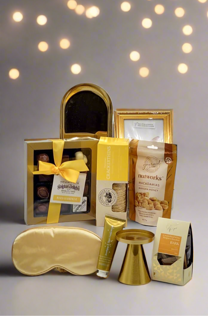 Gifting set with gold sleep mask, chocolate and snacks on a gray background with blurred lights.