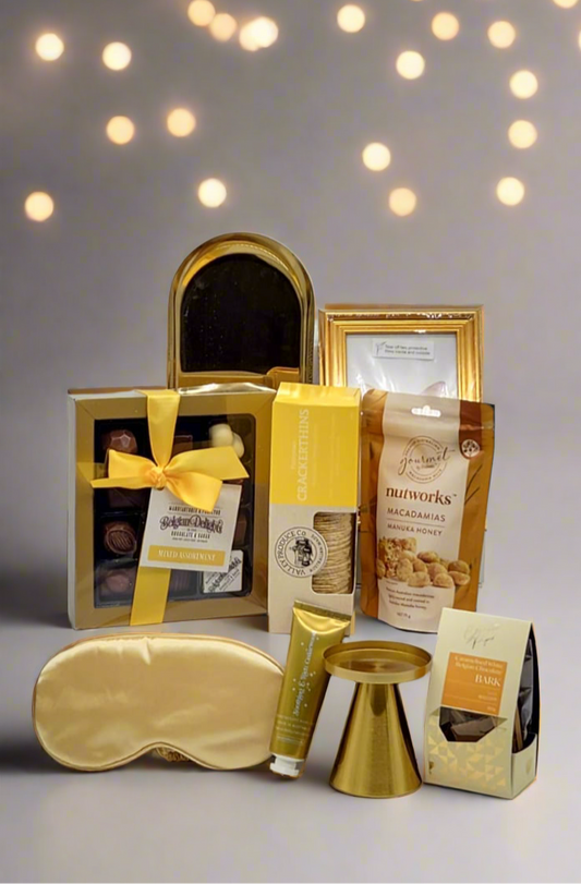 Gifting set with gold sleep mask, chocolate and snacks on a gray background with blurred lights.